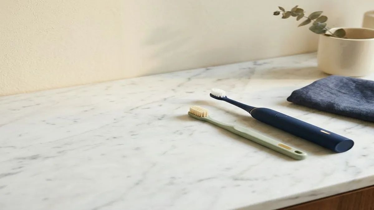 A light green manual toothbrush and a black electric toothbrush side by side on a white marble surface.
