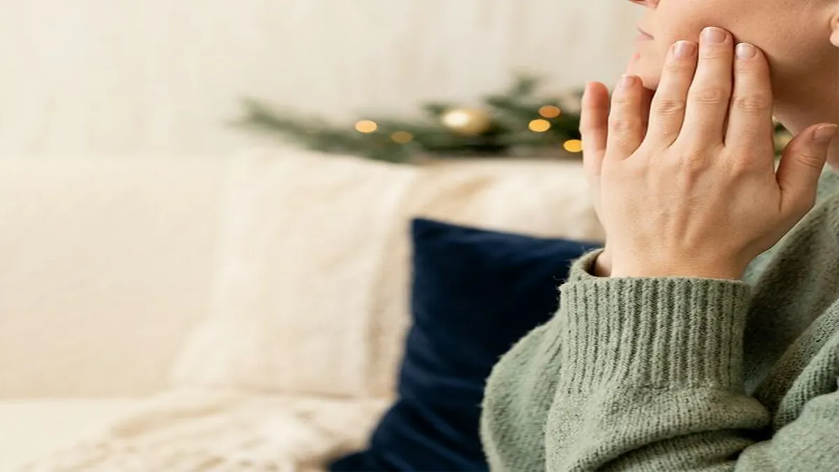 Person in profile with hand to jaw, wearing a sage knit sweater, with blurred evergreen foliage and golden lights in the background.