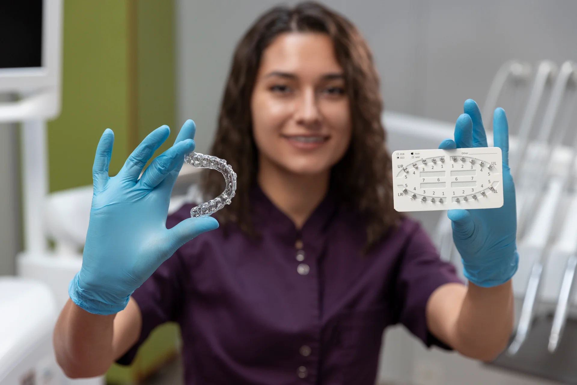 Dental professional in blue gloves holds a clear aligner tray and tooth shade guide in a clinical office setting with a green-tinted wall.
