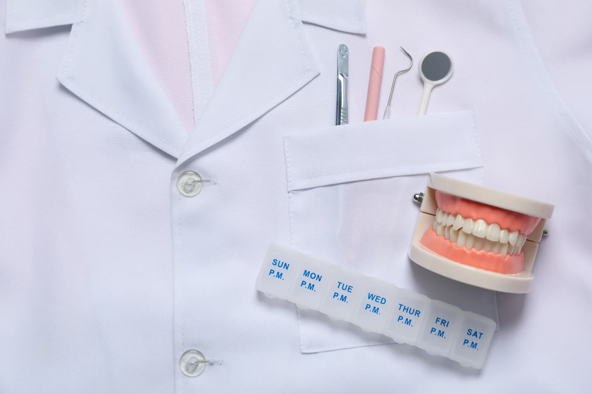 A dental model showing teeth positioned next to a weekly pill organizer and healthcare provider's white coat, representing dental care planning and treatment.