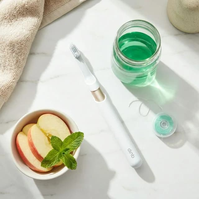 Toothbrush, green mouthwash, sliced apple with mint, and cloth arranged on marble surface.