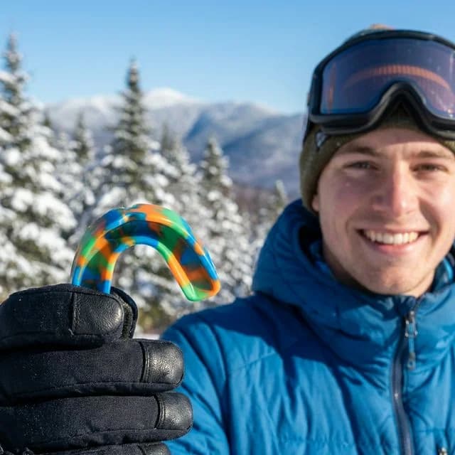 Smiling skier in blue jacket holding a multicolor sports mouthguard with snow-covered mountains and evergreen trees in background.
