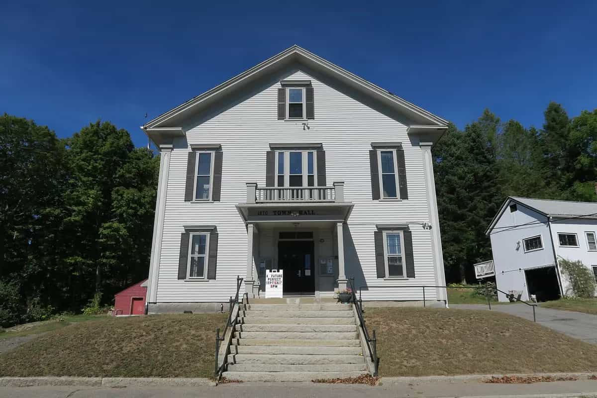 The historic white 1870 Berlin Town Hall in Berlin, Massachusetts, a classic New England clapboard civic building with black shutters and a front portico, under a clear blue sky