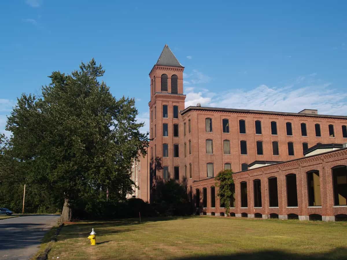 The historic Lancaster Mills brick building with its tall clock tower, a mature shade tree, and green lawn under a clear blue sky in Clinton, Massachusetts