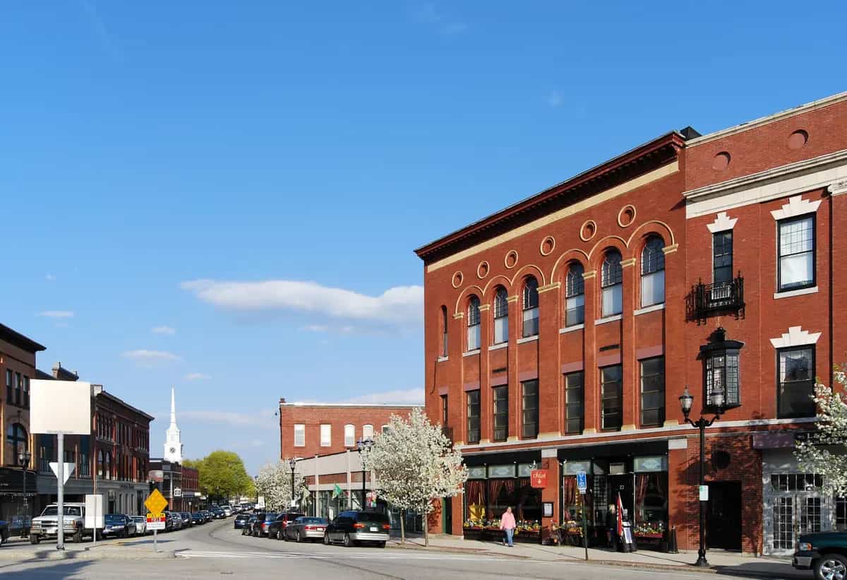 Downtown Main Street in Hudson, Massachusetts on a sunny day, with preserved 19th-century red-brick storefronts, a white New England church steeple in the distance, and blooming spring trees