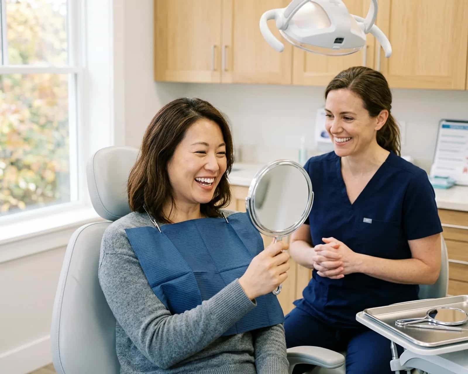 Woman in a dental chair smiling at her reflection in a hand mirror while a clinician looks on