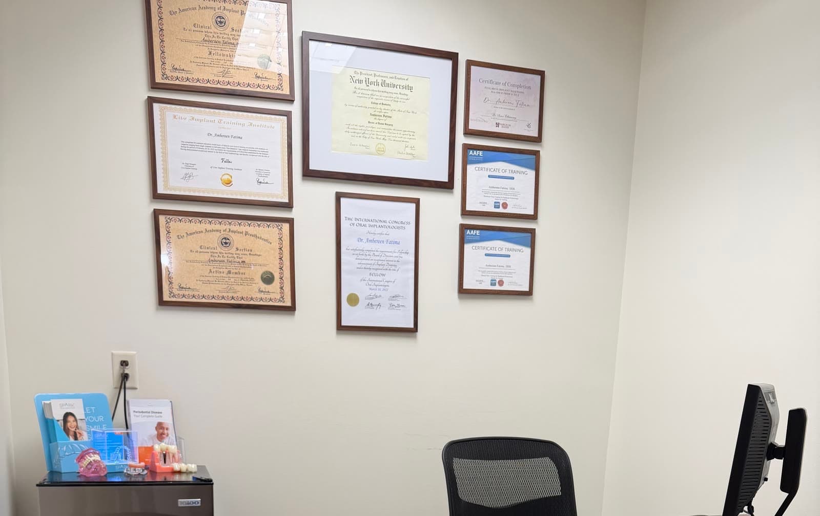 The certificate wall in Dr. Ambereen Fatima's office: a Fellowship and an Active Member certificate from the American Academy of Implant Prosthodontics Clinical Section, a Fellowship from the Live Implant Training Institute, her New York University Doctor of Dental Surgery diploma, a Fellowship from the International Congress of Oral Implantologists, a full-arch All-on-X certificate of completion, and two American Academy of Facial Esthetics certificates of training.